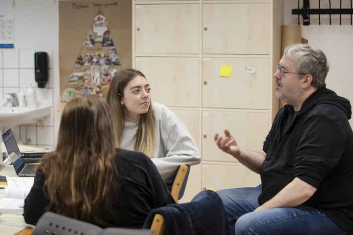 Lærer Tom Akuner sitter og forklarer en oppgave for Emilie og en annen elev med ryggen til i klasserommet.