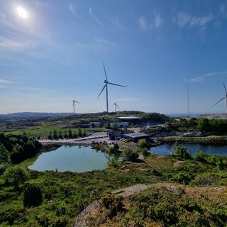 Bilde av området på Svåheia med grønn natur og vann, med vindmøller og blå himmel i bakgrunnen.