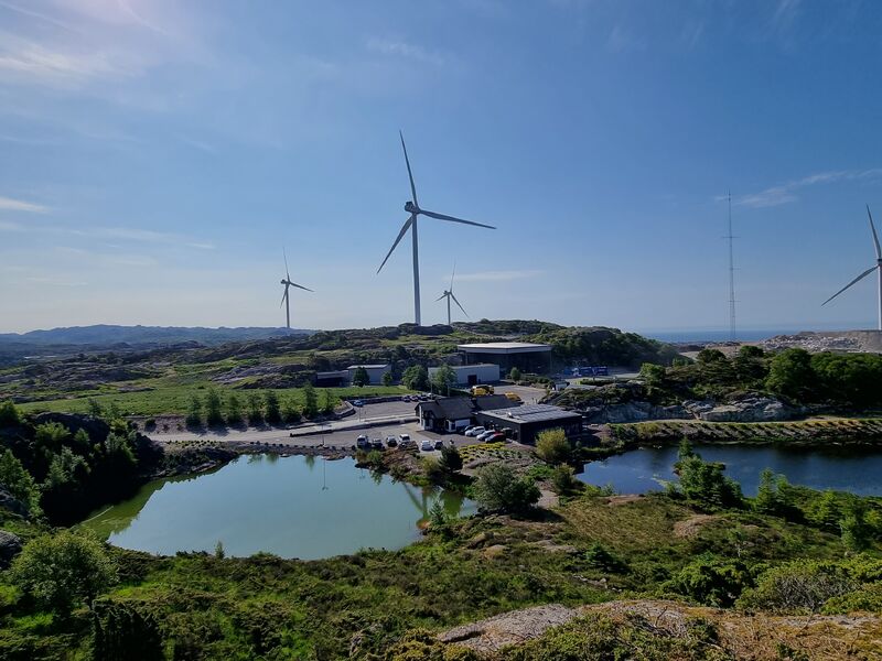 Bilde av området på Svåheia med grønn natur og vann, med vindmøller og blå himmel i bakgrunnen.