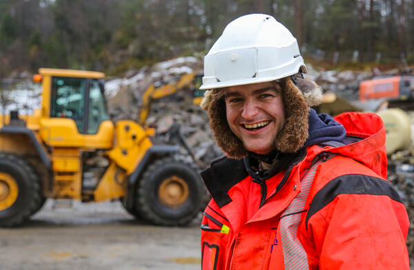 Smilende mann med hvit hjelm står foran en gul gravemaskin