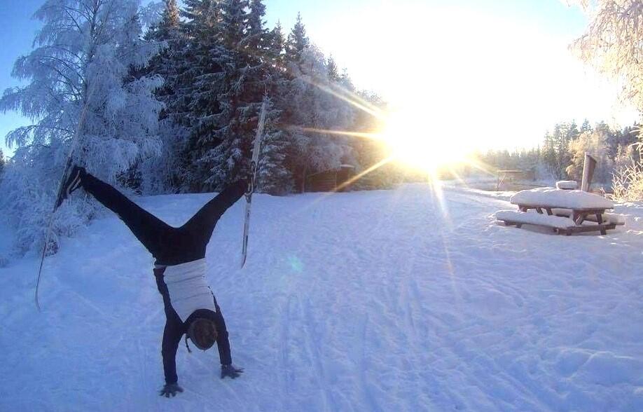 Dame står på hendene i snøen med ski på beina opp mot himmelen