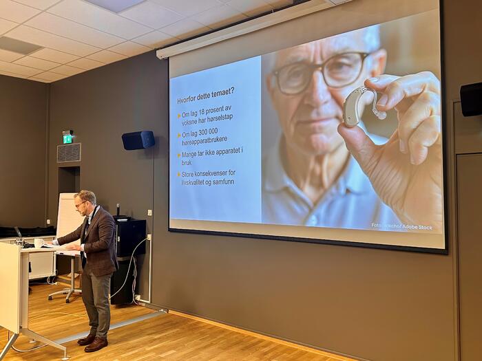 Mann på talerstol med talloversikt i bakgrunnen
