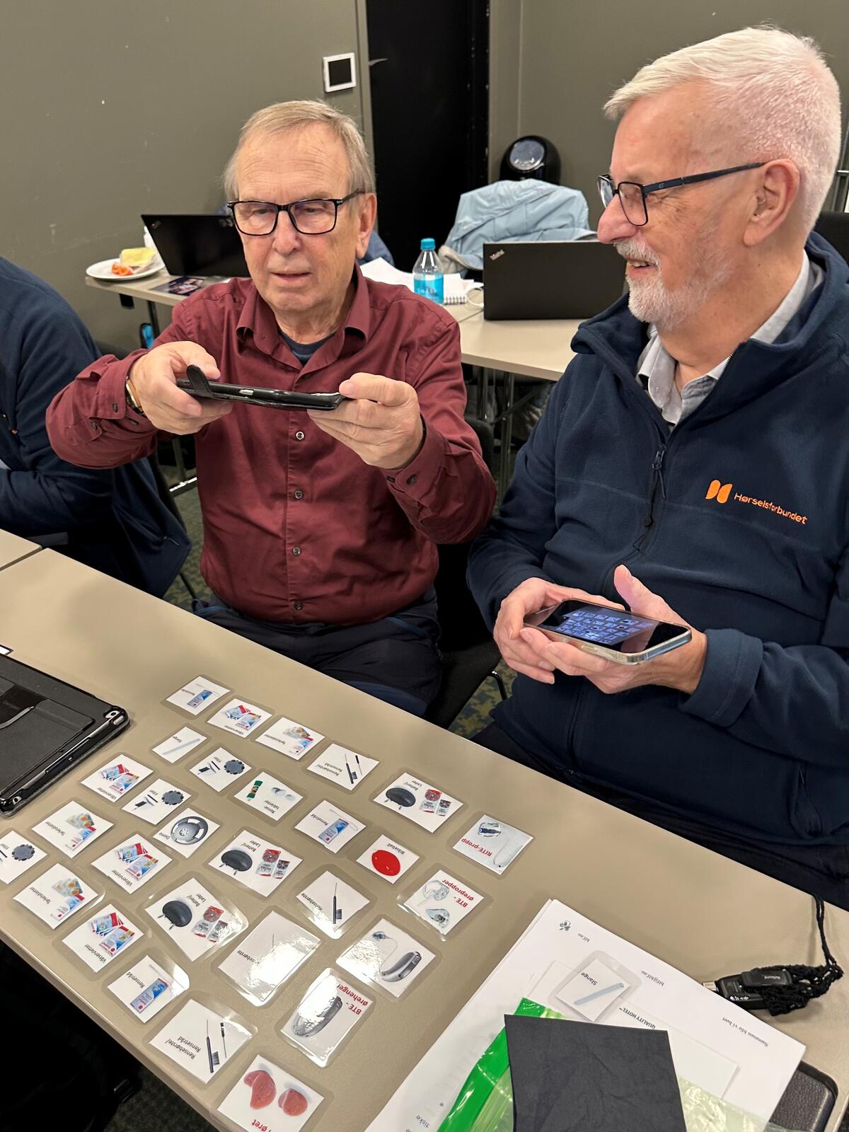 To menn sitter ved bord med kort. En av dem tar bilde med mobilen