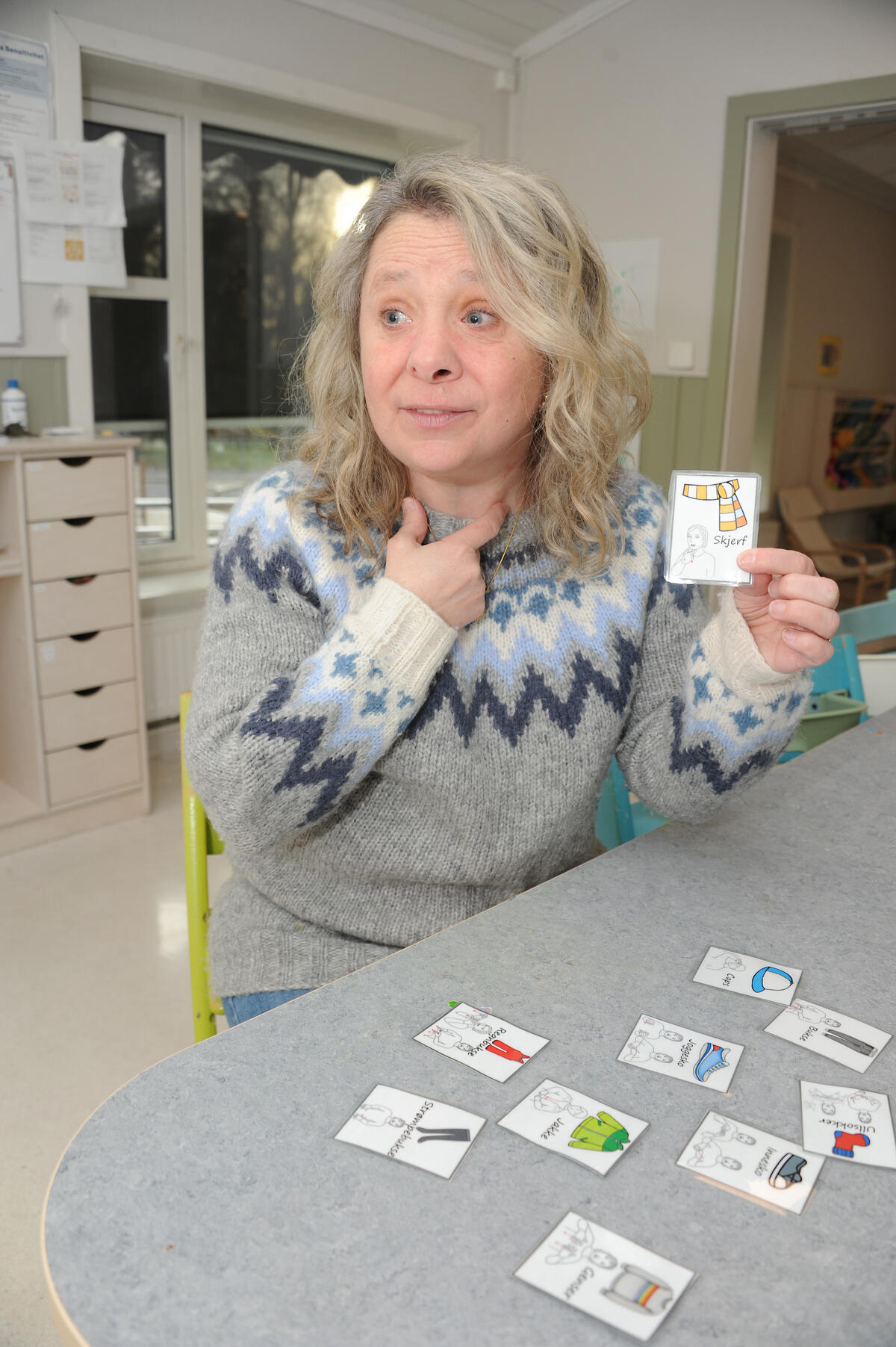 STOR DRØM. Elis store drøm er et alle norske barn skal lære tegn til tale i barnehagen. Hun ser at stadig flere ser nytten av å bruke denne kommunikasjonsformen. Dame i ullgenser sitter med billedkort på et bord og tar seg til halsen