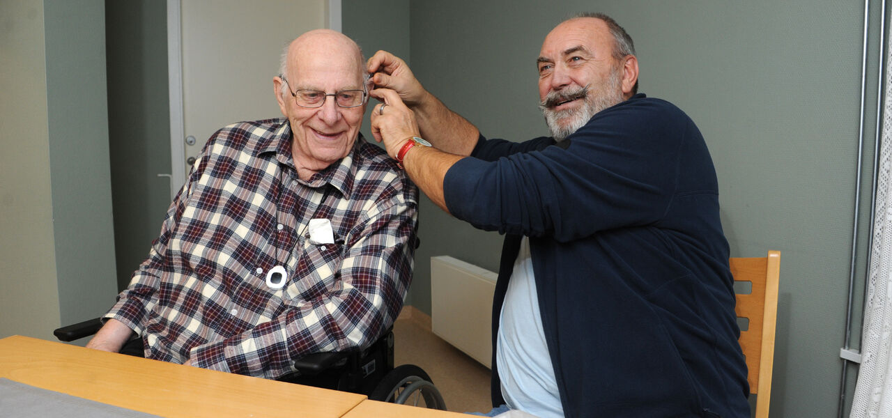 Mann hjelper eldre mann med å sette høreapparat i øret